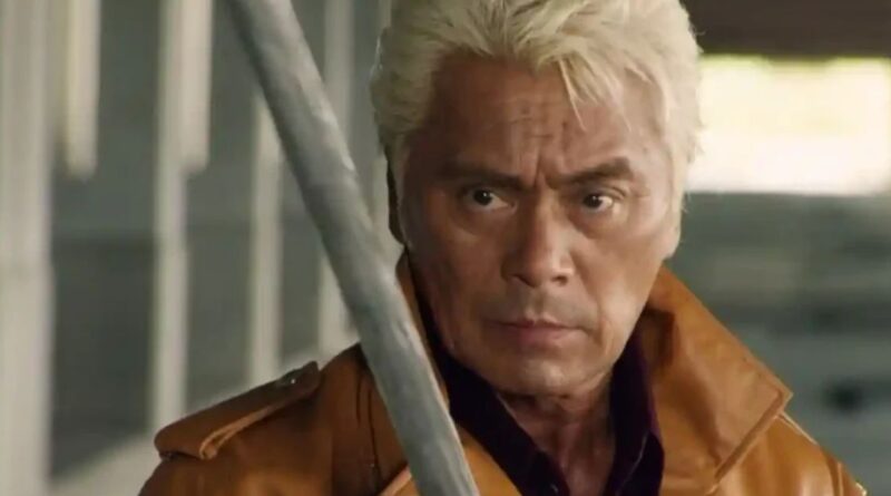 A close-up, dramatic shot of veteran actor Kenji Ohba with blonde hair, wearing a brown leather jacket. He has an intense, focused expression as a silver metallic blade is partially visible in the foreground.