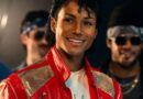 A close-up shot of Jaafar Jackson portraying Michael Jackson, smiling while wearing an iconic red leather jacket with silver sequined shoulders. He is surrounded by other men in 1980s-style clothing and sunglasses in a dimly lit setting.