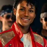A close-up shot of Jaafar Jackson portraying Michael Jackson, smiling while wearing an iconic red leather jacket with silver sequined shoulders. He is surrounded by other men in 1980s-style clothing and sunglasses in a dimly lit setting.