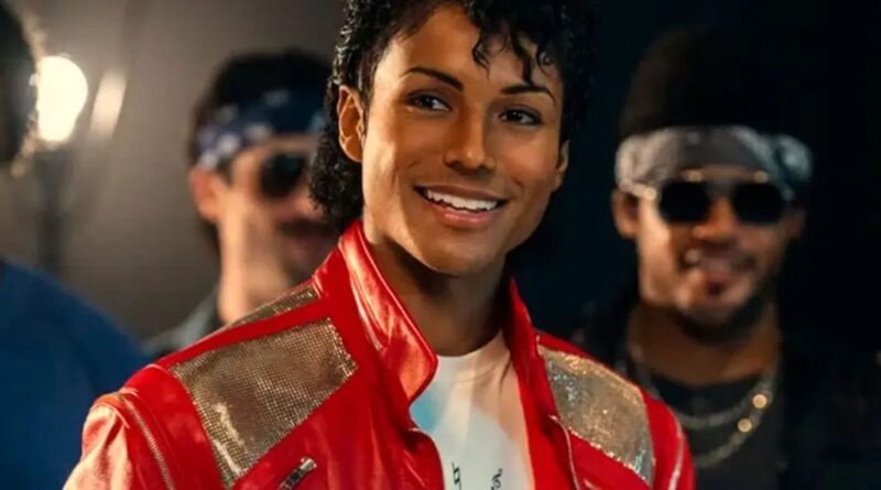 A close-up shot of Jaafar Jackson portraying Michael Jackson, smiling while wearing an iconic red leather jacket with silver sequined shoulders. He is surrounded by other men in 1980s-style clothing and sunglasses in a dimly lit setting.