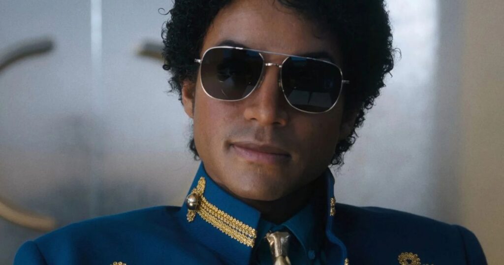 A close-up portrait of Jaafar Jackson portraying Michael Jackson, wearing dark aviator sunglasses and a royal blue military-style jacket with gold braided embroidery on the collar. He has a subtle smile and curly hair, capturing a sophisticated and iconic look from the pop star's career.