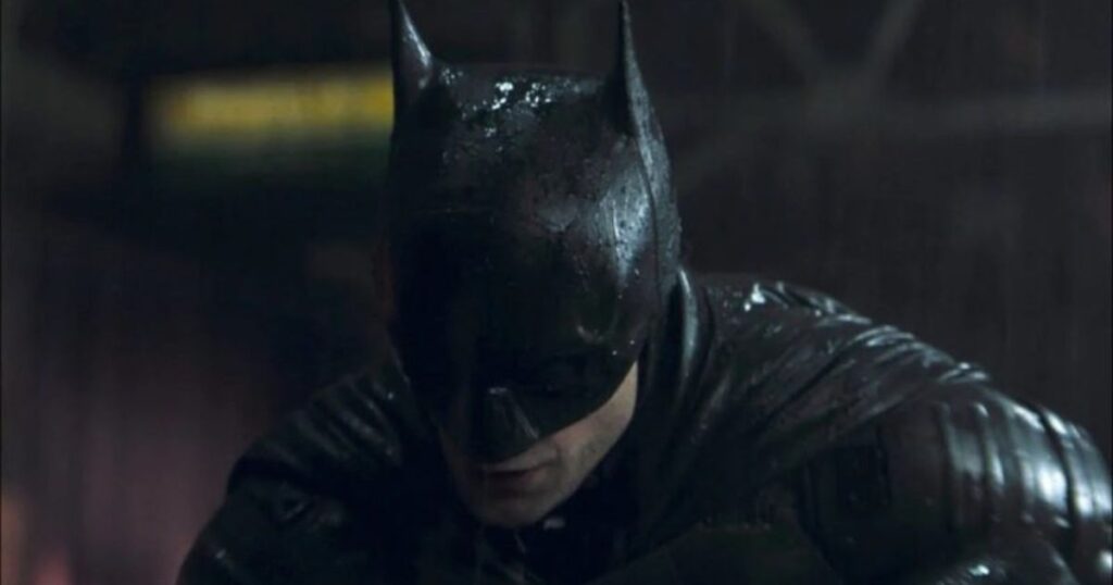 A close-up, dramatic shot of Robert Pattinson as Batman, wearing a wet, tactical cowl under the rain. The lighting is dark and moody, highlighting the texture of the leather-like mask and the character's intense, downward gaze.