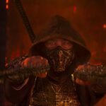 A cinematic close-up of the character Scorpion from Mortal Kombat 2, wearing his iconic gold-detailed mask and dark hood, holding his signature kunai chains forward in a dark, fire-lit environment.