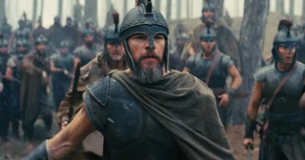 A medium shot of Matt Damon as a Greek warrior, wearing a weathered metal helmet and dark period-accurate armor with a draped cloak. He has a focused, intense expression with a graying beard, leading a group of soldiers through a forest setting. The soldiers in the background are also clad in ancient armor and helmets, creating a gritty and realistic atmosphere.