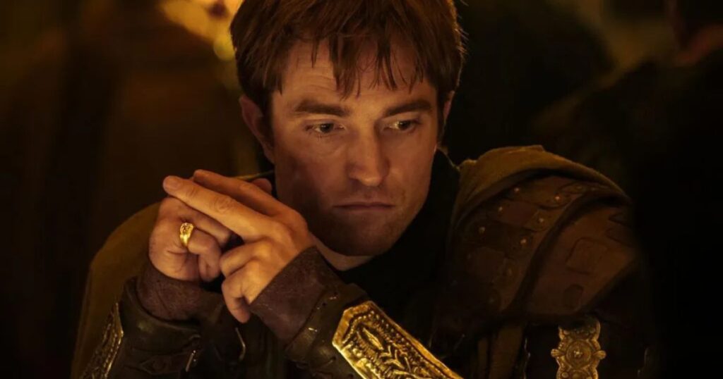 A close-up of actor Robert Pattinson in character for a period piece, potentially for the film 'The Odyssey.' He displays a thoughtful and serious expression, wearing detailed leather armor with gold ornaments on his forearm and a gold ring on his finger, captured under warm, dramatic lighting.