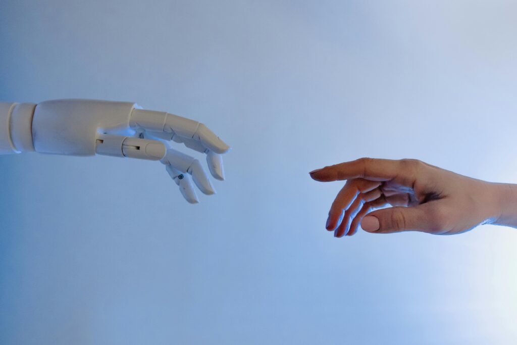 A close-up photograph, with bluish side lighting, showing a white, articulated robotic hand on the left and a human hand on the right, approaching each other without touching. They are positioned horizontally against a light-blue gradient background, reminiscent of Michelangelo's famous fresco 'The Creation of Adam