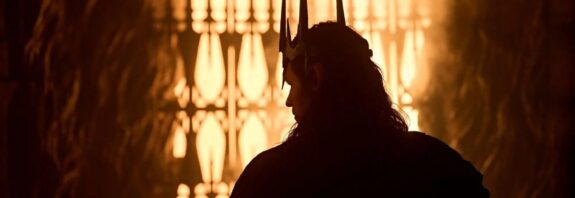 Silhouette of Charlie Vickers as Sauron wearing a spiked crown, standing before a glowing, fiery gate in a scene from The Rings of Power Season 3.
