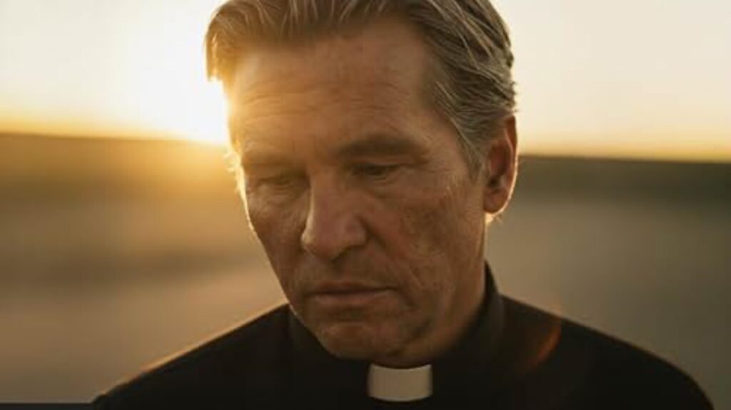 A close-up shot of Val Kilmer wearing a black clerical collar, with his head bowed and a somber expression against a warm sunset.
