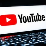 A smartphone lying horizontally on a glowing laptop keyboard, displaying the YouTube logo and wordmark on a white screen against a dark background.