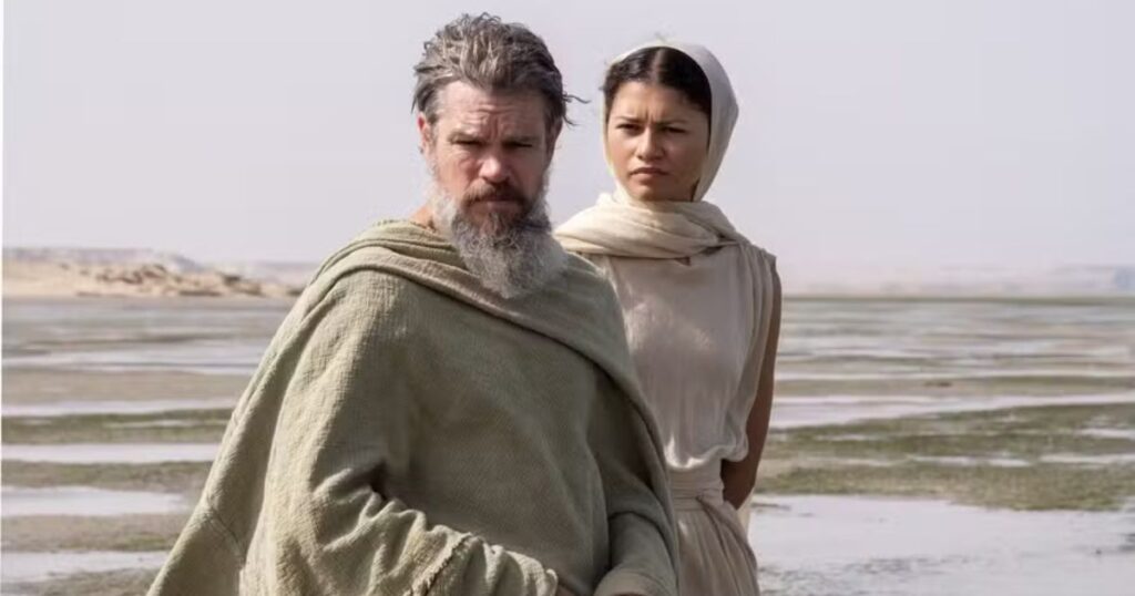 A medium shot of a weathered man with a long gray beard (portrayed by Matt Damon) and a woman with a headscarf (portrayed by Zendaya) standing in a desolate, sandy landscape. Both are dressed in simple, light-colored ancient robes. The man is in the foreground with a weary expression, while the woman stands slightly behind him, looking off into the distance under a bright, overcast sky.